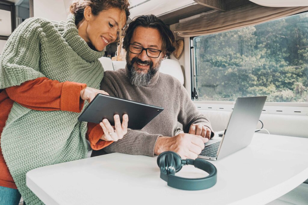 Couple at a table inside an RV. The man sits in front of a laptop and smiles at what the woman is pointing to on her tablet.