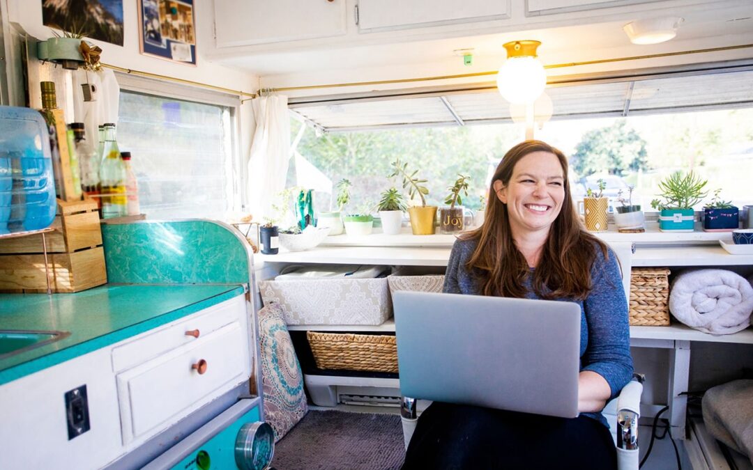 Smiling woman working on a laptop in a colorful and brightly lit RV with several plants in the large window behind her.